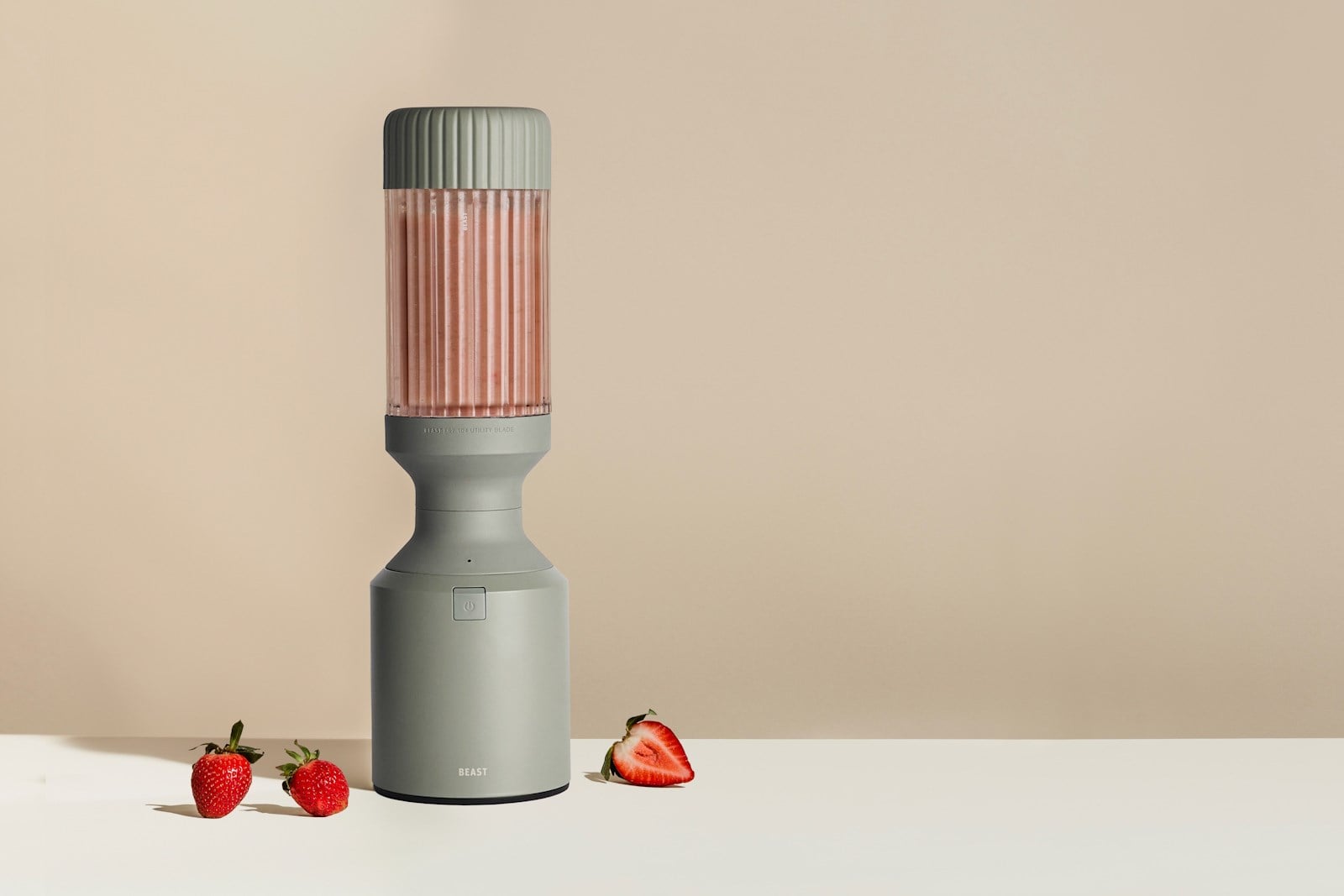 Blender Beast w kolorze Sage z żłobkowanym kielichem napełnionym różowym koktajlem truskawkowym stoi na jasnym blacie, obok całych i przekrojonej truskawki.