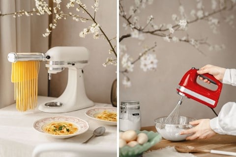 Różowy mikser planetarny KitchenAid z podnoszoną dzieżą na kuchennym stole obok chleba, truskawek i wazonu z kwiatami.